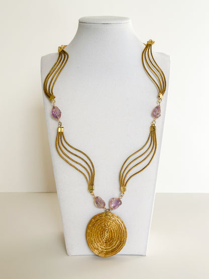 Lua Dourada golden grass medallion necklace featuring handwoven circular pendant with violet crystal accents and matching earrings.