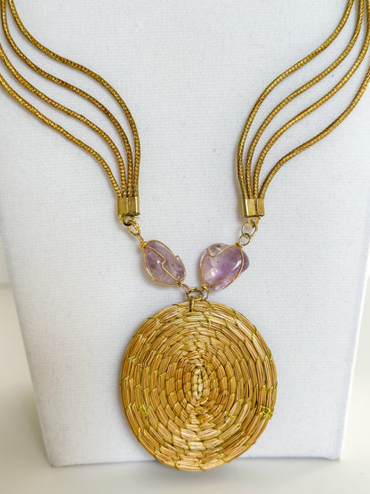 Lua Dourada golden grass medallion necklace featuring handwoven circular pendant with violet crystal accents and matching earrings. close-up
