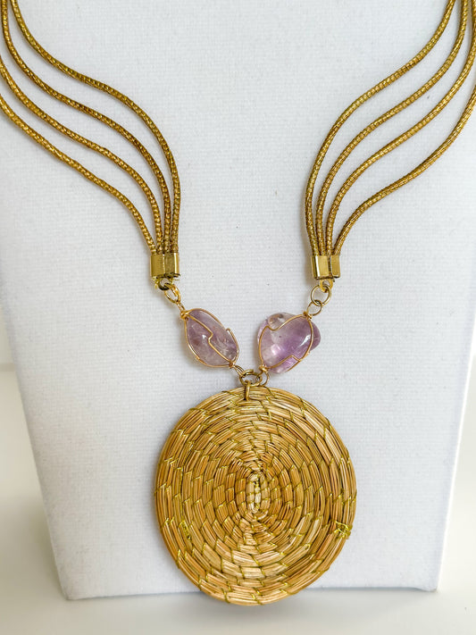 Lua Dourada golden grass medallion necklace featuring handwoven circular pendant with violet crystal accents and matching earrings. close-up