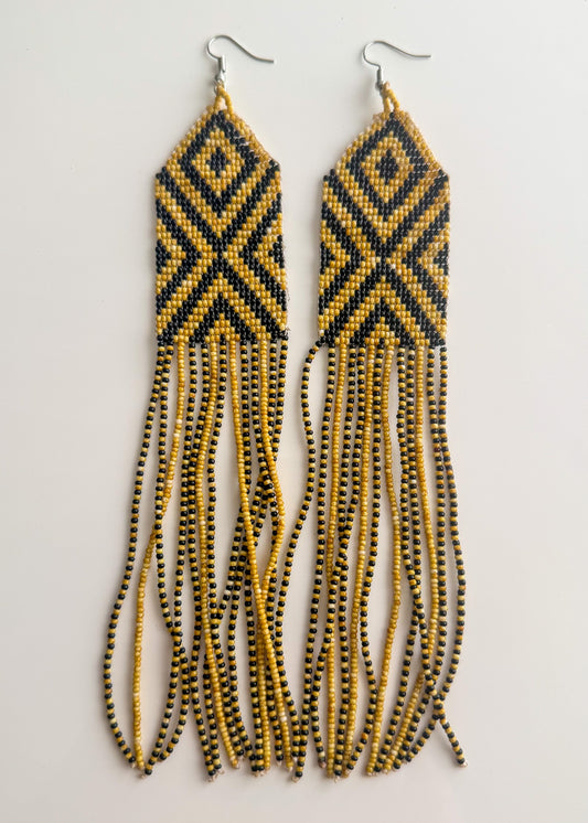 A pair of long beaded dangle earrings with a black and gold geometric diamond pattern on the upper section and long strands of small black and gold beads hanging below, shown against a plain light background.