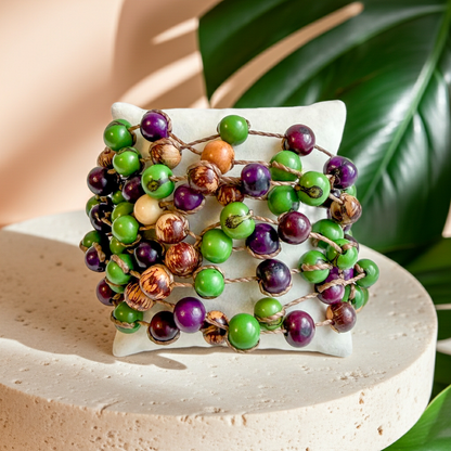 A colorful-acai-seed-statement bracelet with green, purple, and brown beads, displayed on a small white cushion atop a round beige stone pedestal with tropical leaves in the background.
