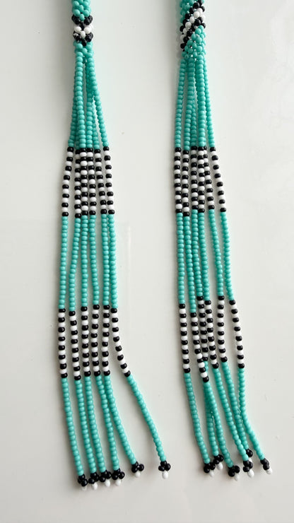 A pair of long beaded fringe necklaces or tassels made of turquoise, black, and white beads, displayed against a plain white background.