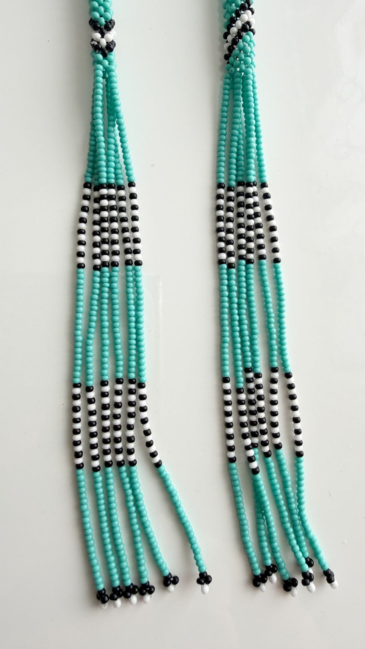 A pair of long beaded fringe necklaces or tassels made of turquoise, black, and white beads, displayed against a plain white background.